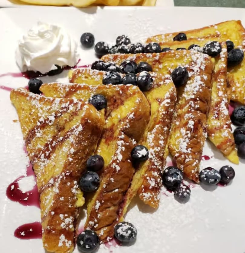 Golden French toast slices topped with blueberries, powdered sugar, and whipped cream on a white plate.