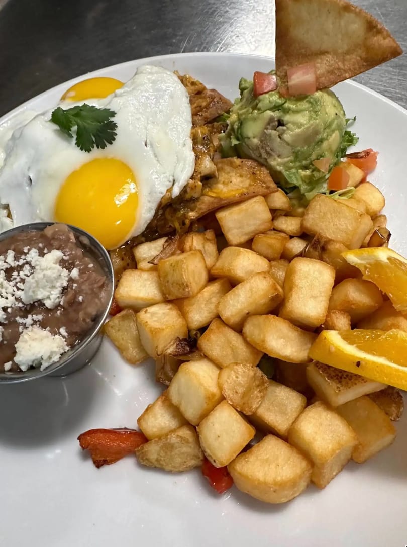 A breakfast plate with two fried eggs, home fries, refried beans, guacamole, and a tortilla chip.