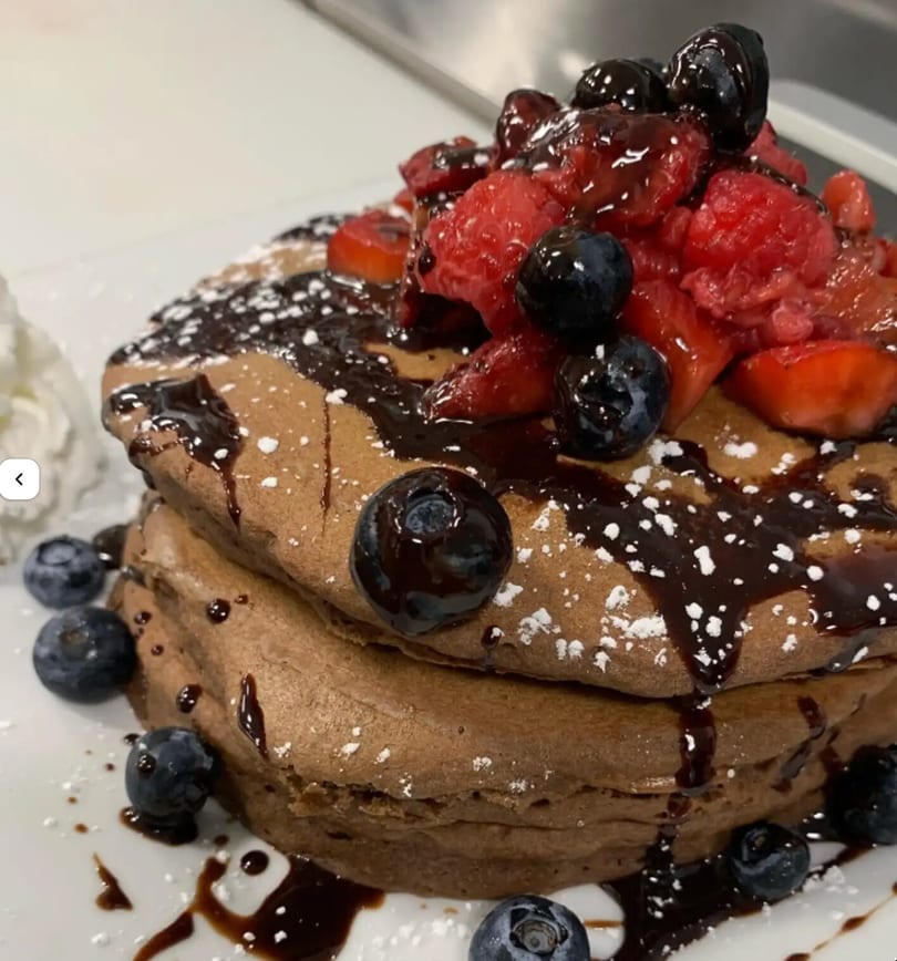Stack of chocolate pancakes topped with fresh berries, chocolate syrup, and powdered sugar.