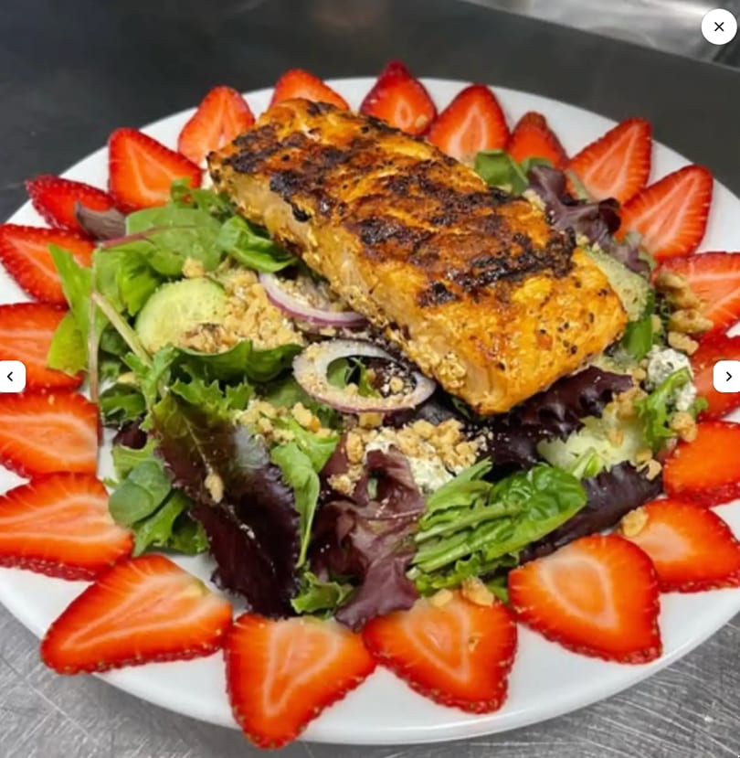 Grilled salmon fillet on a bed of mixed greens, topped with nuts and surrounded by strawberry slices.